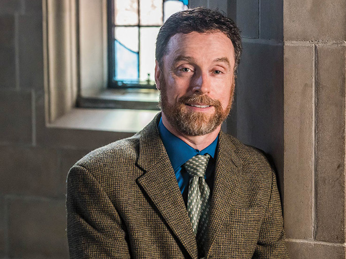 A man with short dark hair and a beard, wearing a brown tweed jacket, blue shirt, and patterned tie, smiles with a sense of progress while standing by a stone wall with a window in the background.