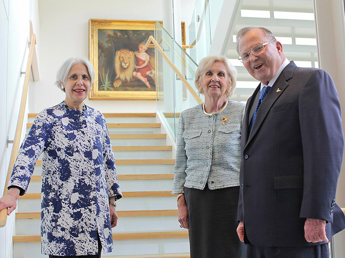 Three people stand smiling at the base of a staircase, exuding warmth and empathy. A painting of a child with a lion graces the wall behind them. The individuals include two women and a man, all dressed formally, showcasing their empathy skills through genuine expressions.