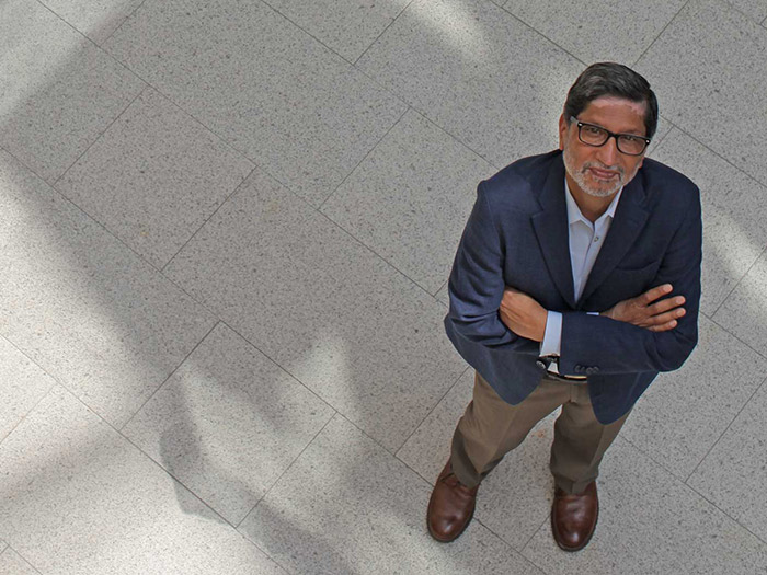 A man with glasses, donning a navy blazer and brown shoes, stands with arms crossed, gazing toward the future. He is on a light gray tiled floor at the university, with dynamic shadows elegantly cast across it.