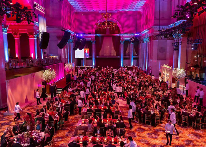 A large, elegant banquet hall filled with people seated at round tables, adorned with red and white floral centerpieces. The room is illuminated with warm, colorful lighting. Waitstaff in white shirts move efficiently between tables, ensuring guests receive exceptional service—a true return on investment in memorable experiences.