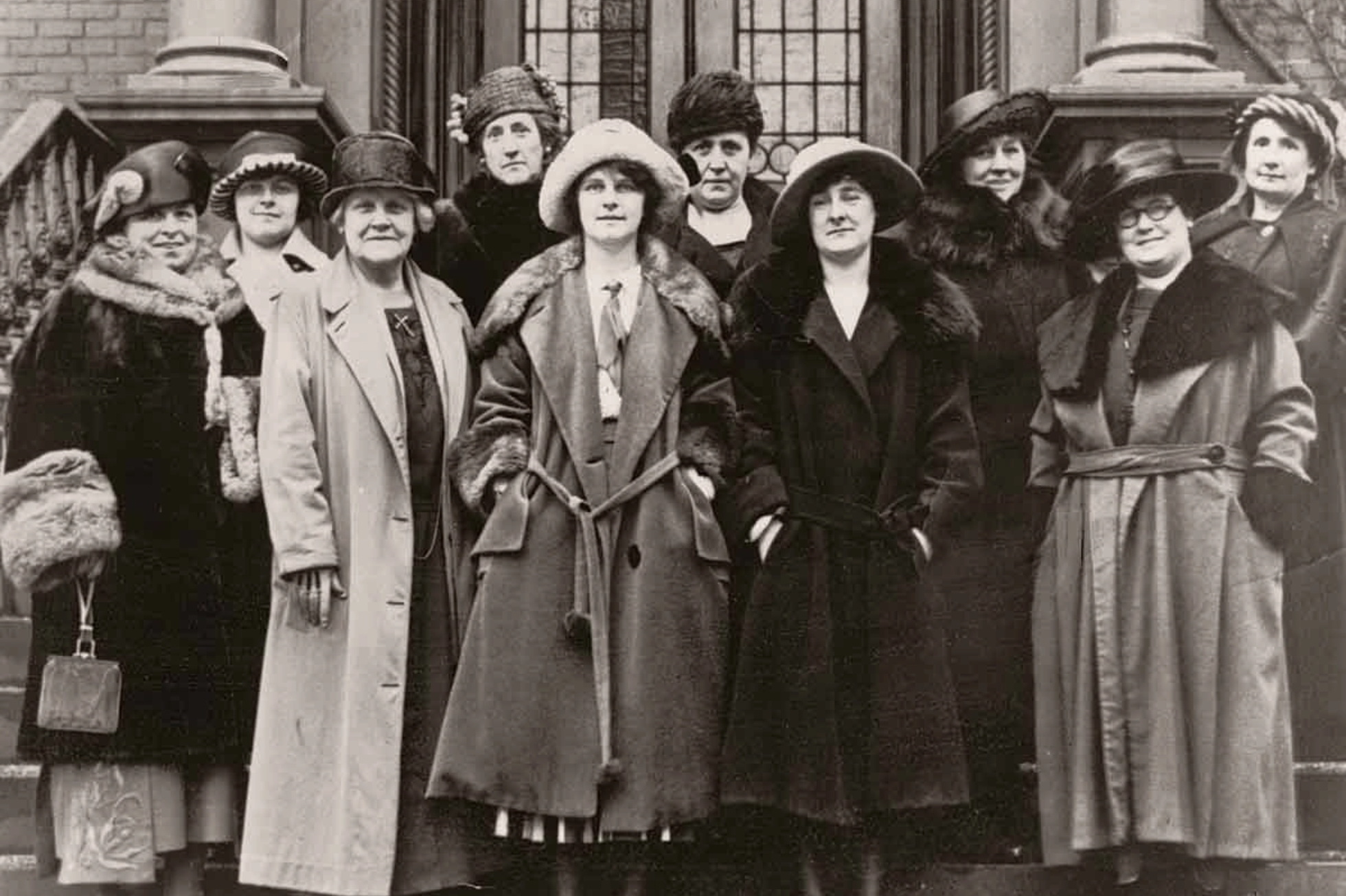 A group of ten women from the early 20th century stand in front of a building. They are wearing long coats and various styles of hats, some with fur collars, raising their heights. The background features ornate brickwork and a stained glass window.