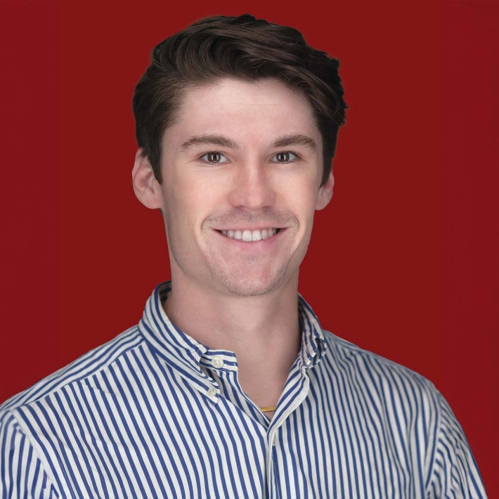 A person with short brown hair smiling, wearing a blue and white striped shirt. The background is solid red.