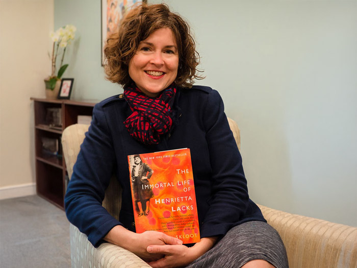 A person with curly hair is sitting in a chair, smiling while holding "The Immortal Life of Henrietta Lacks." Clad in a dark jacket and patterned scarf, they reflect on the Lacks legacy. A small bookshelf and a potted plant sit calmly in the background, echoing stories yet to be told.