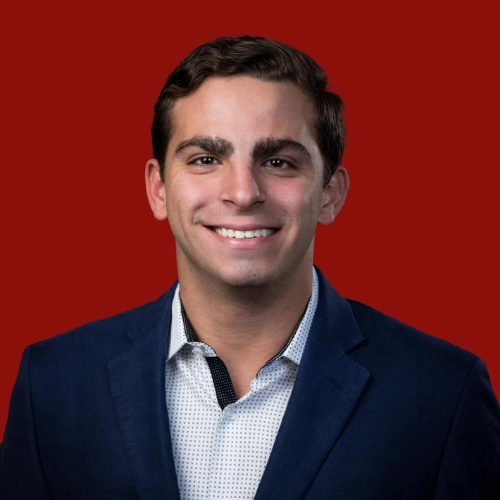Smiling person in a blue blazer and dotted shirt against a solid red background.
