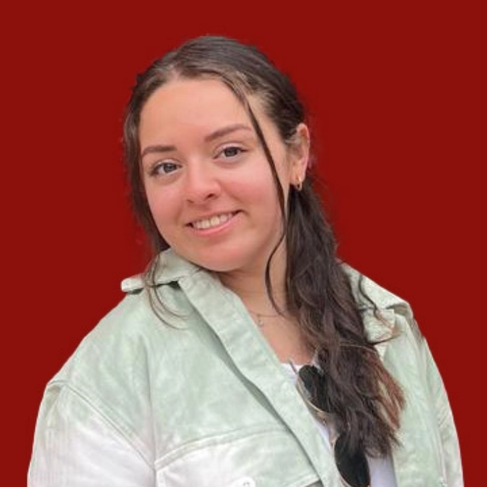 A person with long dark hair smiles at the camera, wearing a light-colored jacket over a white top. They have sunglasses hanging from the collar. The background is solid red.