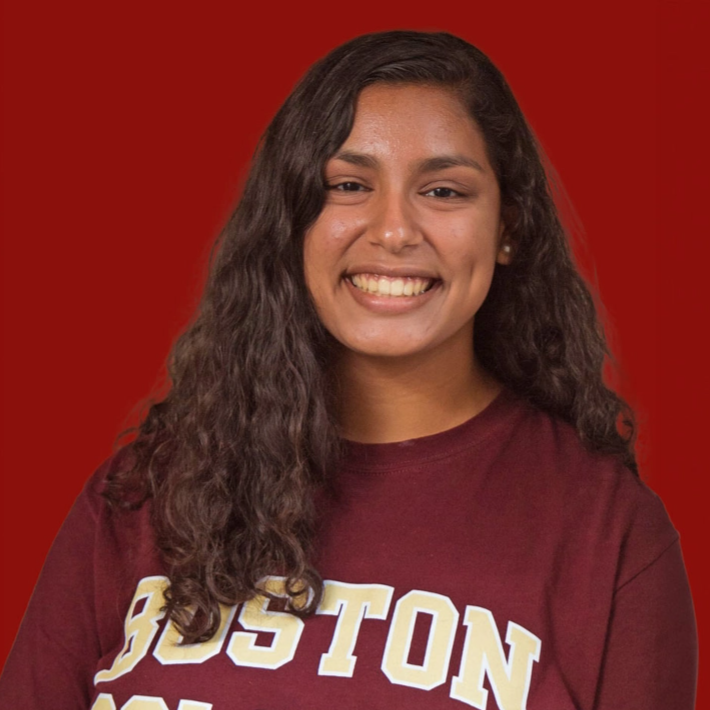A person with long curly hair and a wide smile is wearing a maroon Boston College shirt. The background is solid red.