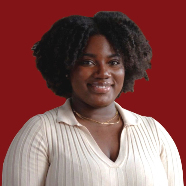 A person with curly hair wearing a cream-colored top smiles while facing forward. The background is a solid maroon color.