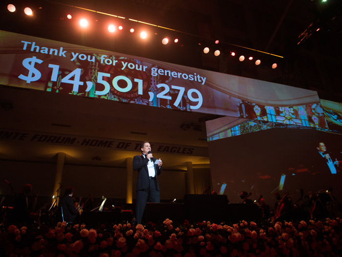 A person stands on stage holding a microphone, hitting a high note, as the large screen behind them displays "Thank you for your generosity $14,501,279." Red flowers line the stage and an orchestra is visible in the background. The atmosphere of this performance is celebratory.