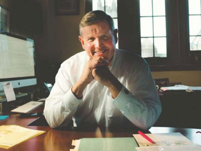 A smiling person sits with persistence at a desk in an office, wearing a white shirt. Papers and a red pen are scattered as if in an irrational arrangement. A computer waits in the background while sunlight filters through windows, crafting a warm atmosphere.