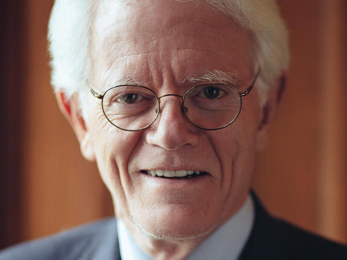Elderly man with white hair and glasses smiling warmly, wearing a suit and tie—a true embodiment of the gift that still gives. The background is softly lit.