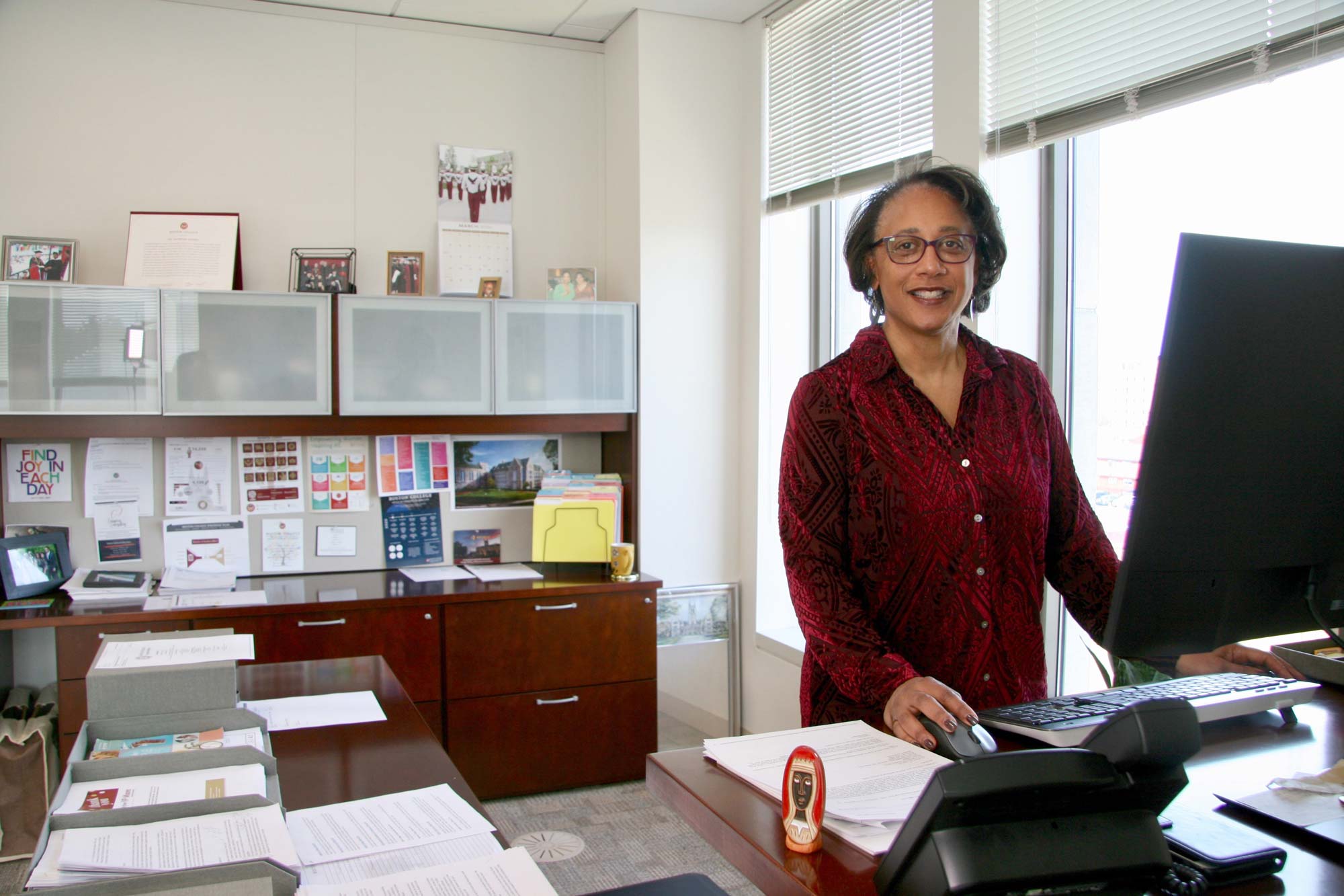 A woman stands smiling at a desk with a computer in a bright, organized office. The room features file cabinets, framed certificates, and colorful artwork on the walls. Sunlight streams through large windows, creating a welcoming atmosphere.