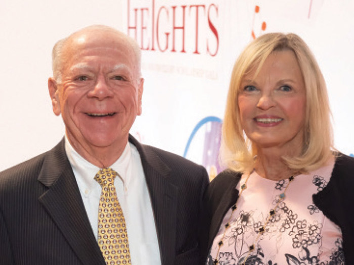 An elderly man and woman are smiling. The man is in a dark suit with a patterned tie, while the woman wears a floral dress and black cardigan. Having reached a significant landmark, they stand before a backdrop with the text "Heights" partially visible.
