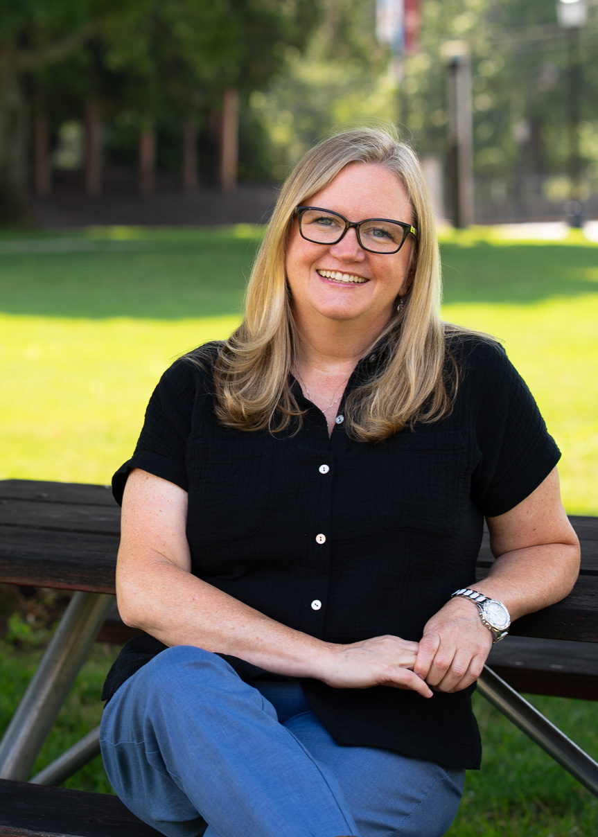 A woman with long blonde hair and glasses, wearing a black short-sleeve shirt and blue pants, sits smiling on a picnic bench in a sunny park with green grass and trees in the background.