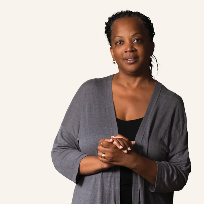 A woman with braided hair stands against a plain light background, wearing a gray cardigan over a black top. Her calm, friendly expression reflects the Cura Universitatis spirit as she gently clasps her hands in front of her.