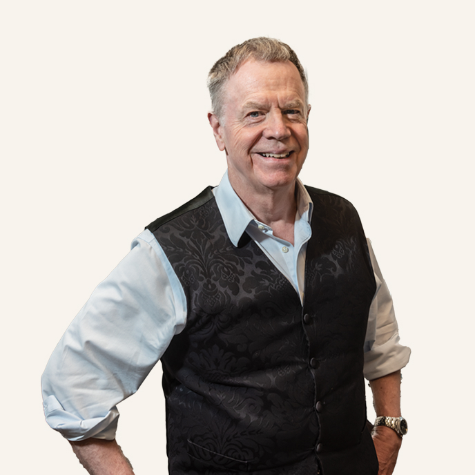 Smiling older man with gray hair wearing a light blue shirt and a black patterned vest, standing with one hand on his hip against a plain light background, embodying the spirit of university tradition.