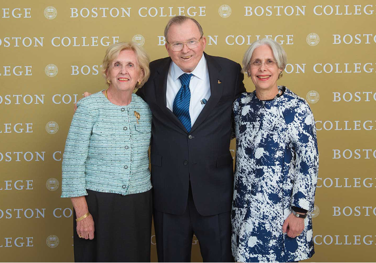 Three older adults, two women and one man, stand smiling in front of a gold Boston College backdrop. Dressed in professional attire, they embody the Cura Universitatis spirit and Winston Tradition as they pose closely together.