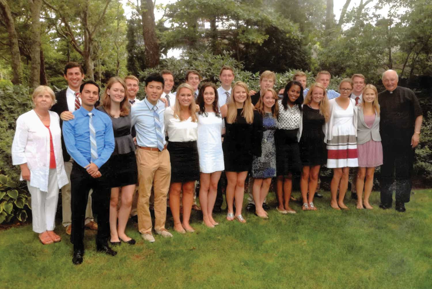 A group of about 24 people, dressed in semi-formal attire, stand closely together outdoors on a grassy lawn with trees and bushes in the background, smiling at the camera as they celebrate a cherished university tradition.