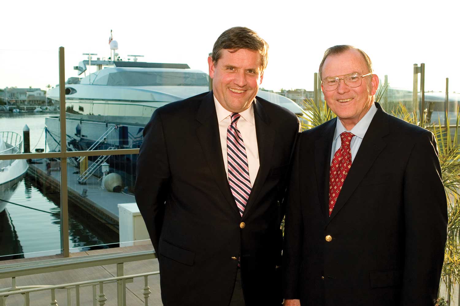 Two men in suits stand smiling on a waterfront balcony with a large yacht and marina in the background, embodying the spirit of Winston Tradition on a sunny day.