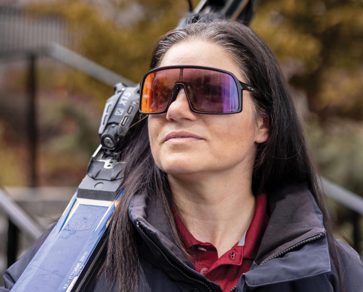 A woman with long dark hair wearing large, reflective sunglasses and a dark jacket carries skis on her shoulder outdoors, with a blurred background of autumn foliage.