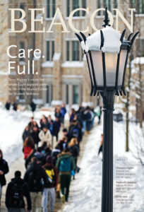 A snow-covered lamppost stands in focus in the foreground, while groups of people bundled in winter clothes walk along a snow-lined campus pathway in the background.