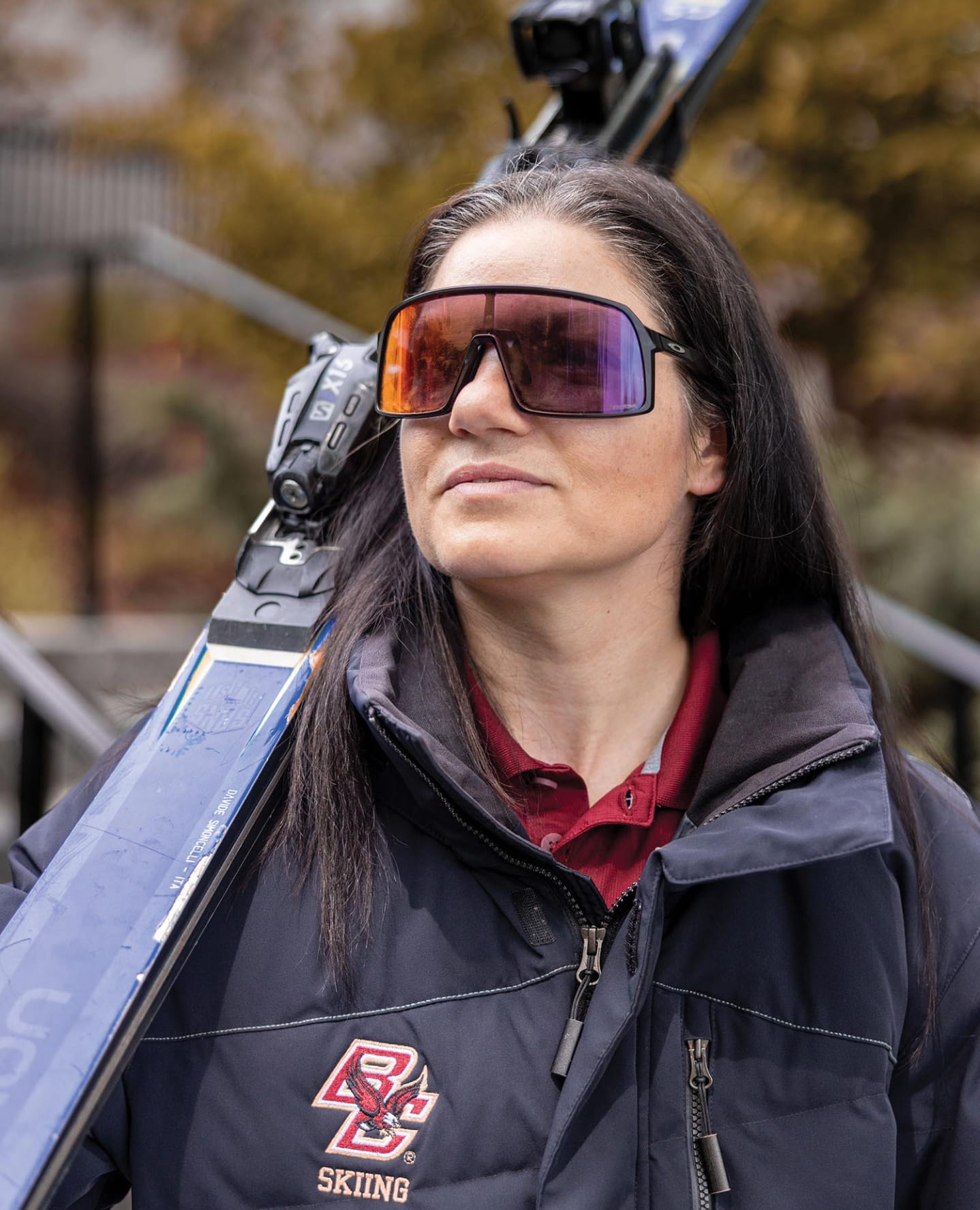 A woman wearing oversized sunglasses and a dark jacket with a "BC Skiing" patch carries skis over her shoulder outdoors, with blurred trees in the background.