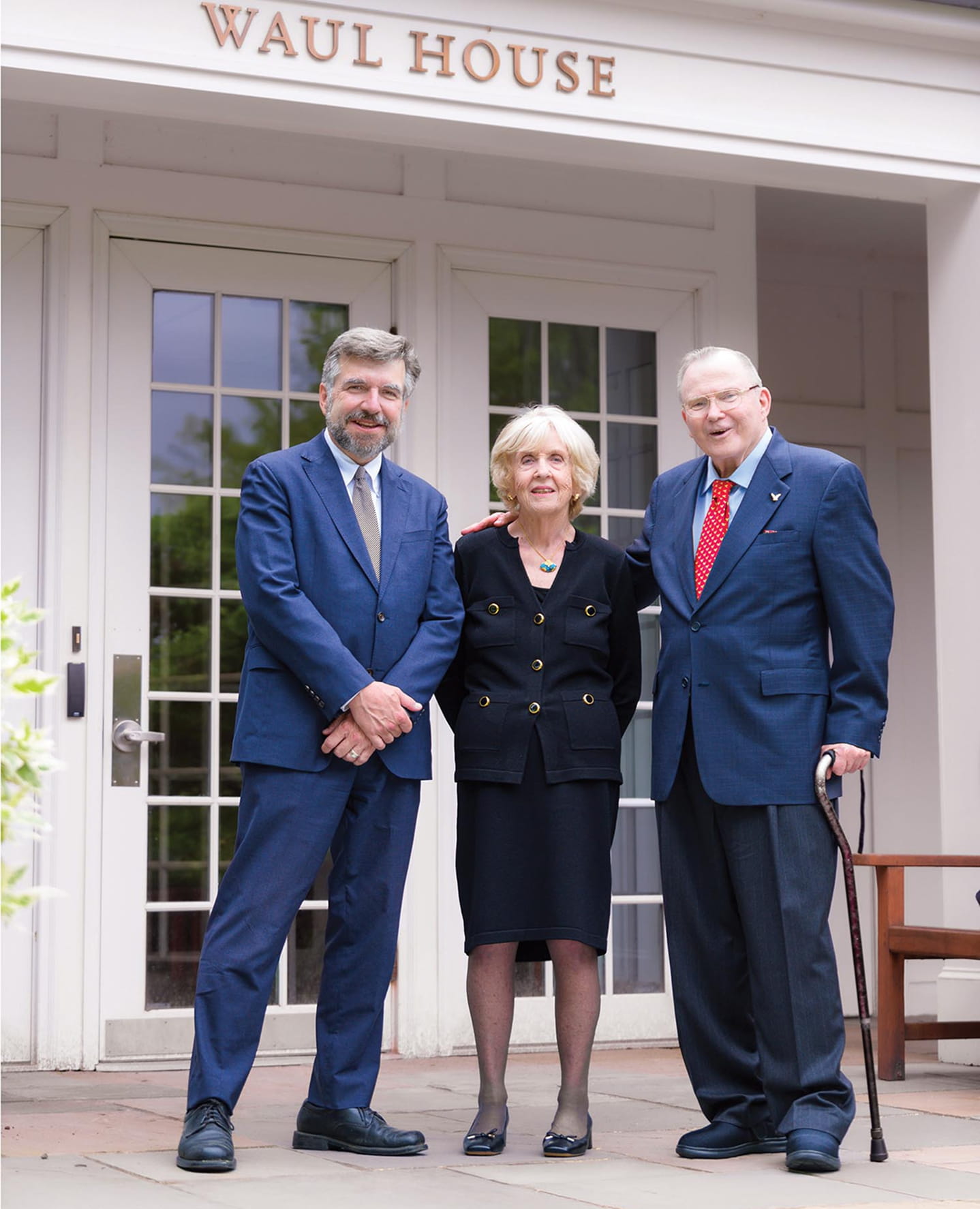 A woman wearing oversized sunglasses and a dark jacket with a "BC Skiing" patch carries skis over her shoulder outdoors, with blurred trees in the background.