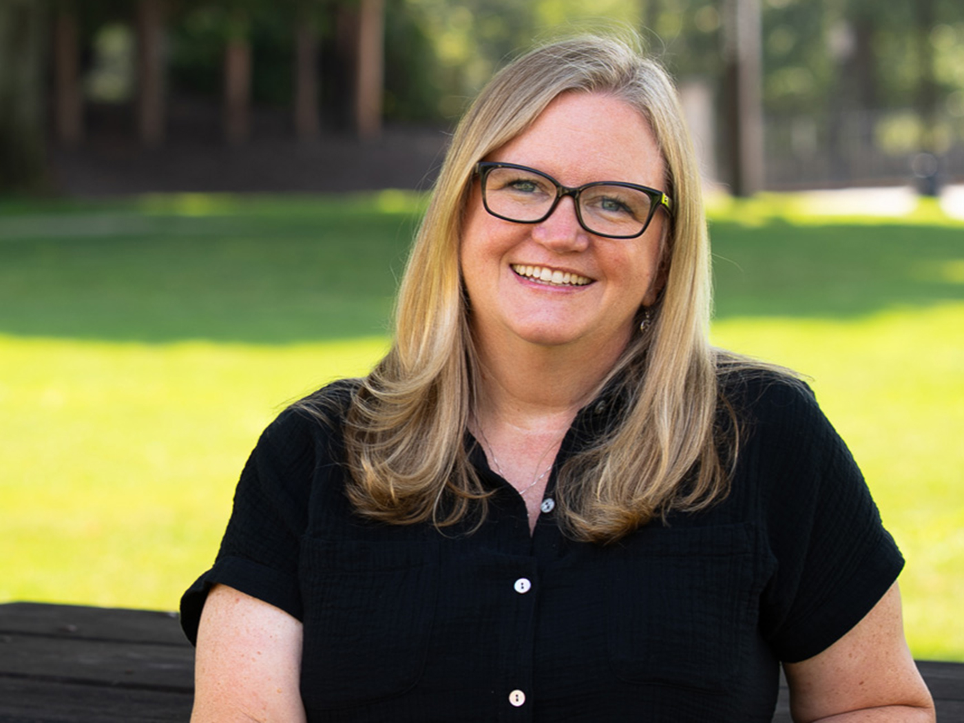 A woman with long blonde hair and glasses, wearing a black shirt, smiles while sitting outdoors on a sunny day with green grass and trees—perfect for a feature in Beacon Magazine.