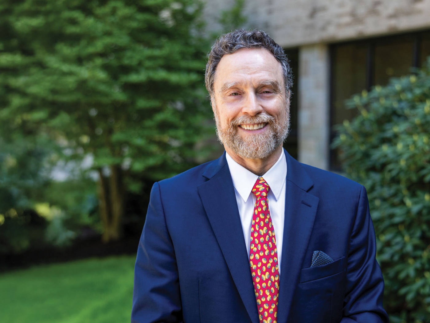 A smiling man with a beard, wearing a navy suit, patterned red tie, and pocket square, stands outdoors in front of greenery and a building.