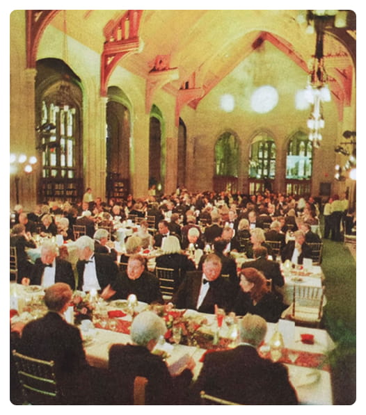 A large group of people dressed formally are seated at long, decorated tables in a grand hall with high arched ceilings and tall windows, attending a banquet or gala event.