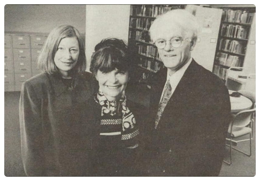 Three adults, two women and one man, stand together indoors, smiling at the camera. Bookshelves, filing cabinets, and a table with chairs are visible in the background. The image is in black and white.