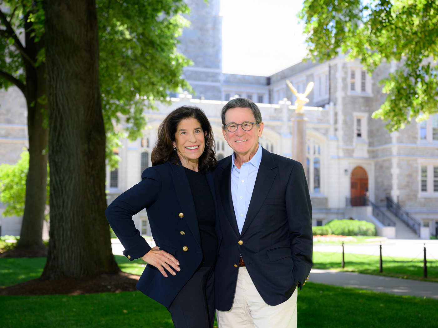 A smiling woman and man in business attire stand together outside on a green lawn, with a large, elegant stone building and leafy trees in the background on a sunny day.
