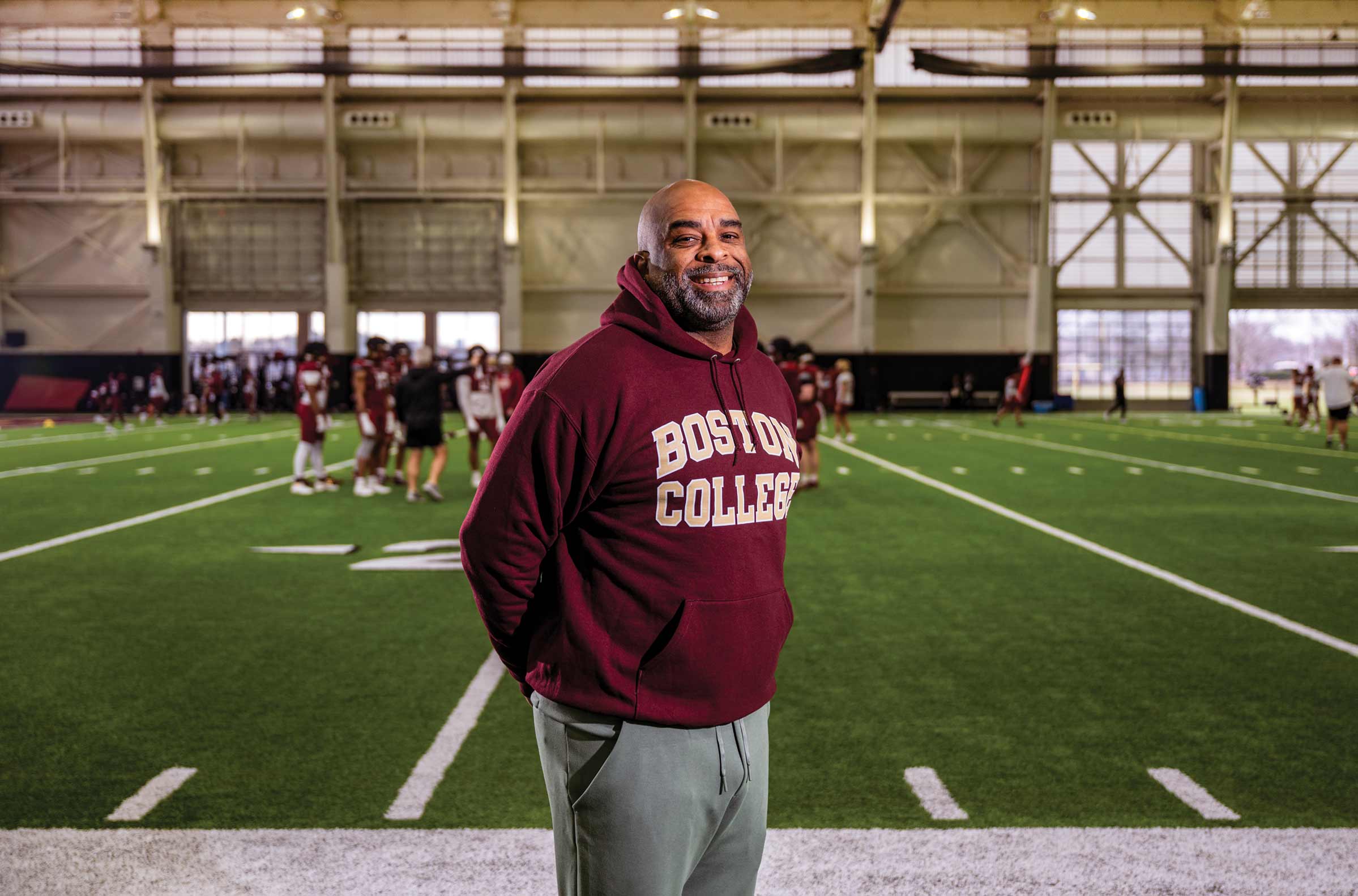 A smiling man in a maroon "Boston College" hoodie stands on an indoor football field, showing his BC Football pride. Several players in maroon uniforms practice in the background, with large windows and bright overhead lights above.
