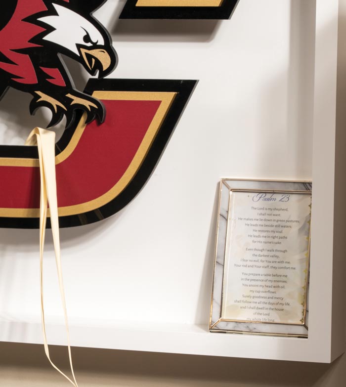 A framed excerpt of Psalm 23 sits on a white shelf next to a large decorative letter "E" featuring a stylized eagle logo in red, black, and gold—a nod to Family First values in BC. A ribbon hangs from the shelf.