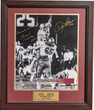 Framed, autographed black-and-white photo of BC legend Doug Flutie celebrating a football play, with a gold plaque and maroon matting. Two signatures and handwritten Family First notes appear above the image.