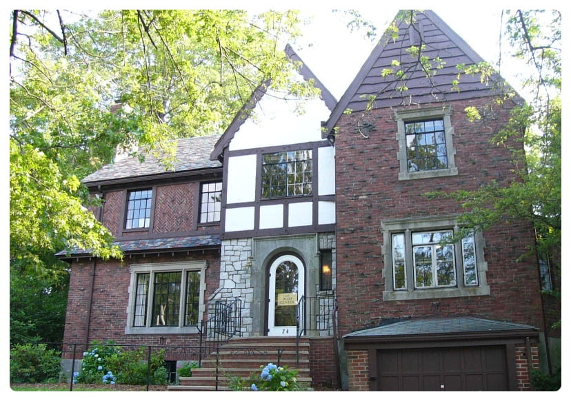 A two-story brick Tudor-style house with decorative half-timbering, large windows, an arched doorway, a front porch with stairs, and a single-car garage, surrounded by trees and greenery.