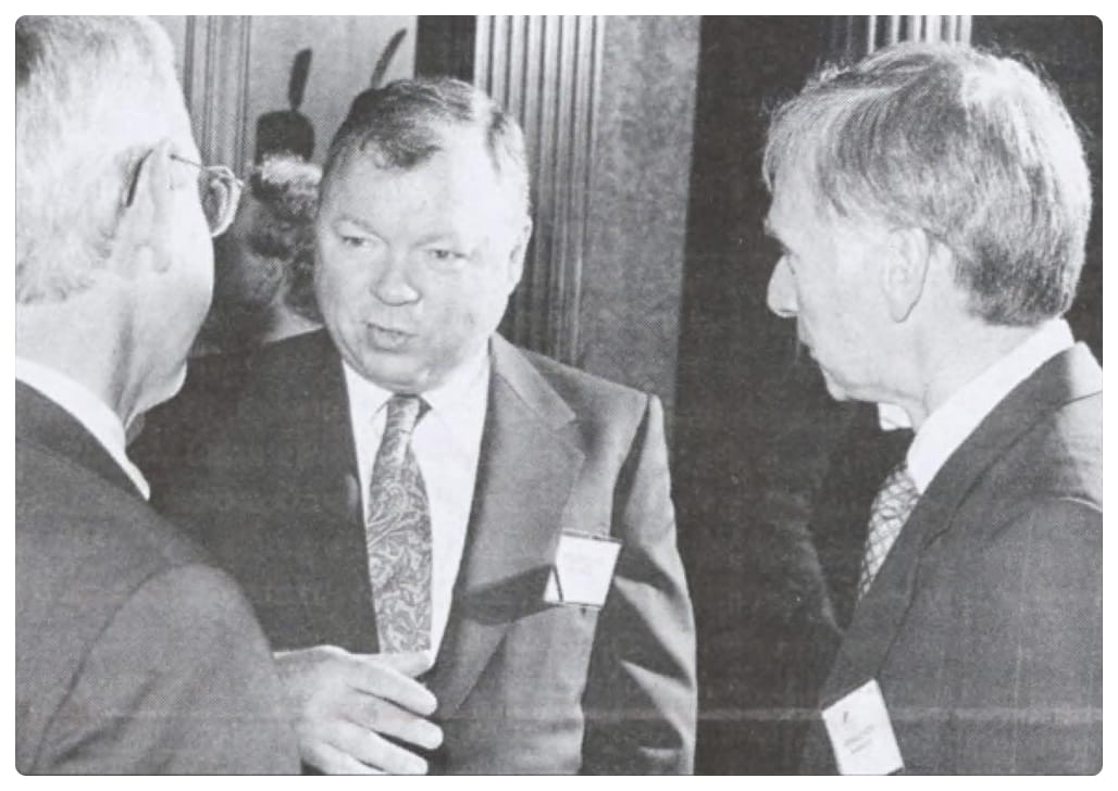Three men in suits and ties are engaged in conversation at a formal indoor event. Two men face a third man who is speaking; all wear name badges. The setting includes tall columns and other people in the background.