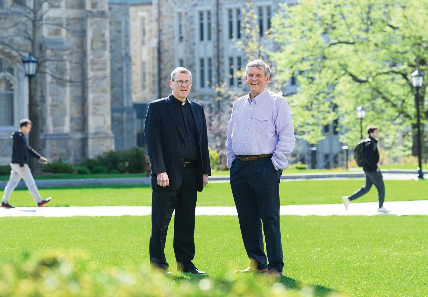 Two men stand smiling on a grassy campus with trees and stone buildings in the background, perfectly capturing a moment of striking the match for new beginnings. Other people walk by on paths in the distance, all set against a bright, sunny day.
