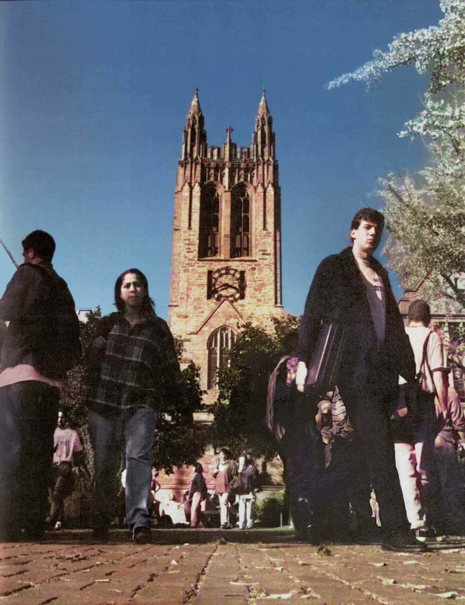 Groups of people walk in front of a tall, stone, gothic-style tower that rises as a steady beacon under a clear blue sky, with trees partially framing the scene.
