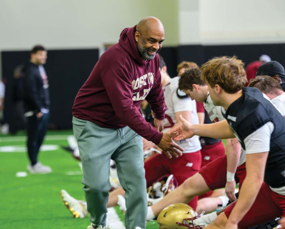 A coach in a maroon hoodie and gray pants enthusiastically greets a football player sitting on the turf, while other players stretch nearby during an indoor practice—capturing the kind of spirited moments often featured in Beacon Magazine.