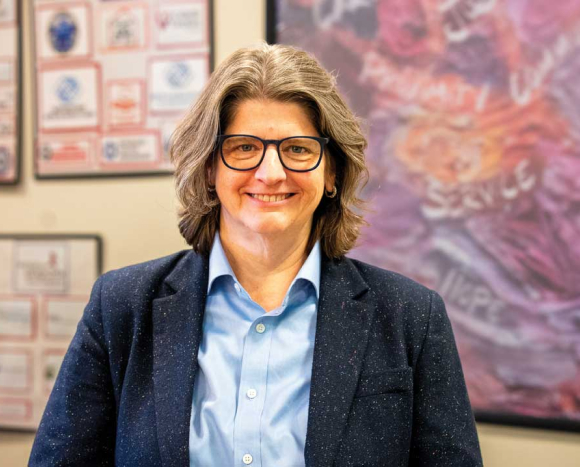 A person with shoulder-length brown hair and glasses, wearing a blue shirt and dark blazer, smiles while standing indoors. Colorful art and framed documents—including a feature from Beacon Magazine—are visible on the wall behind them.