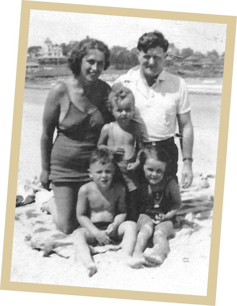 A black-and-white photo of a family of five posing on a beach blanket. Two adults stand behind three young children, with sand and buildings visible in the background, capturing a quiet act of gratitude shared among loved ones at the shore.