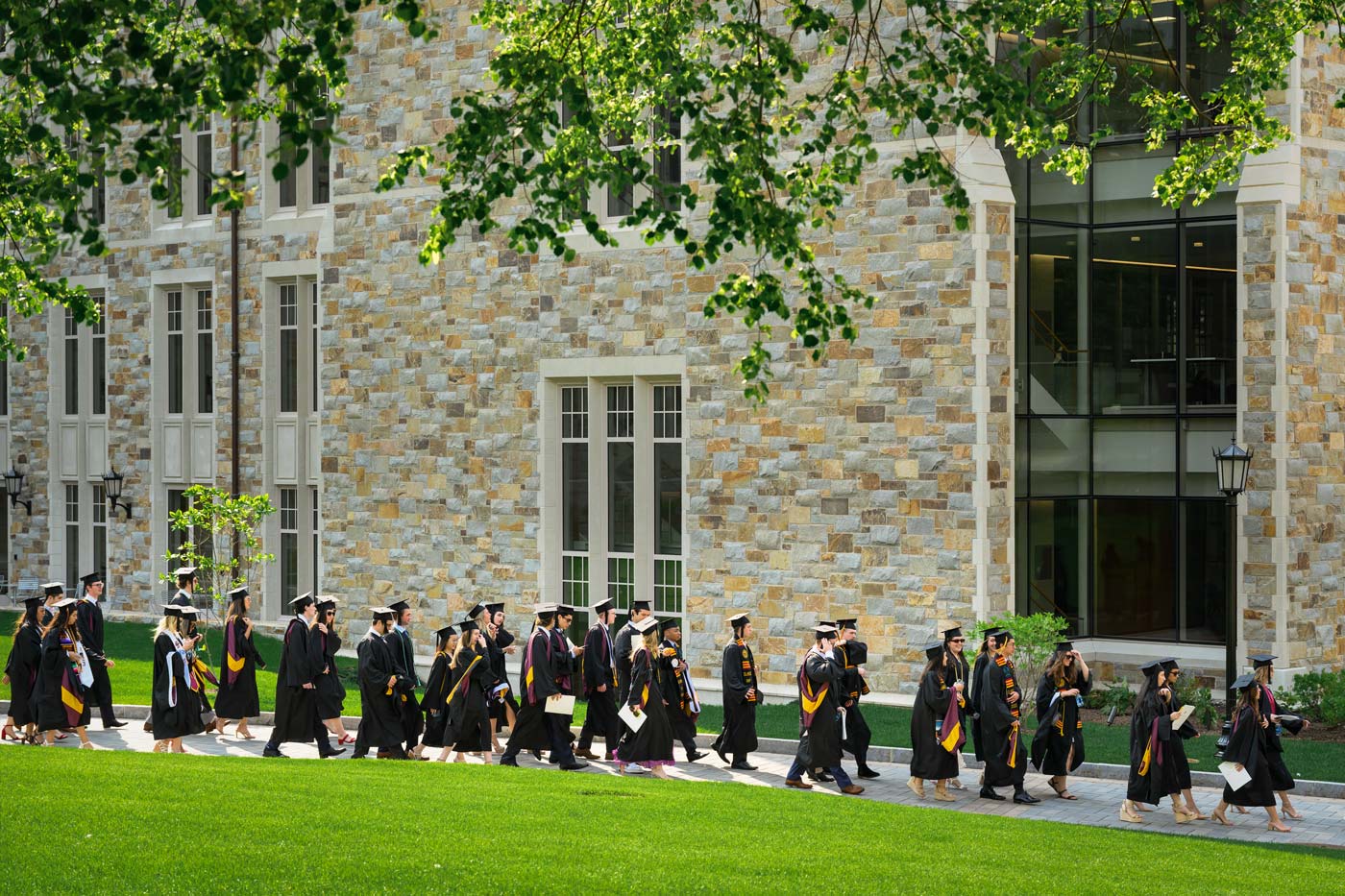 A steady group of graduates in caps and gowns walk in a procession along a path next to a large stone building, surrounded by green grass and trees, their future bright as a beacon on this sunny day.