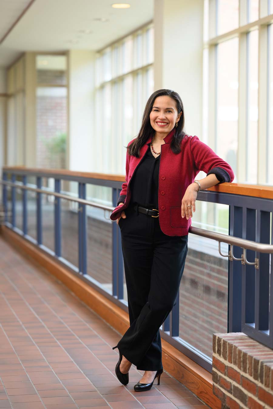 Woman with long dark hair, wearing a red blazer, black top, and black pants, stands smiling in a brightly lit hallway with large windows and a blue railing, striking the match of confidence as she leans against the railing with one hand in her pocket.