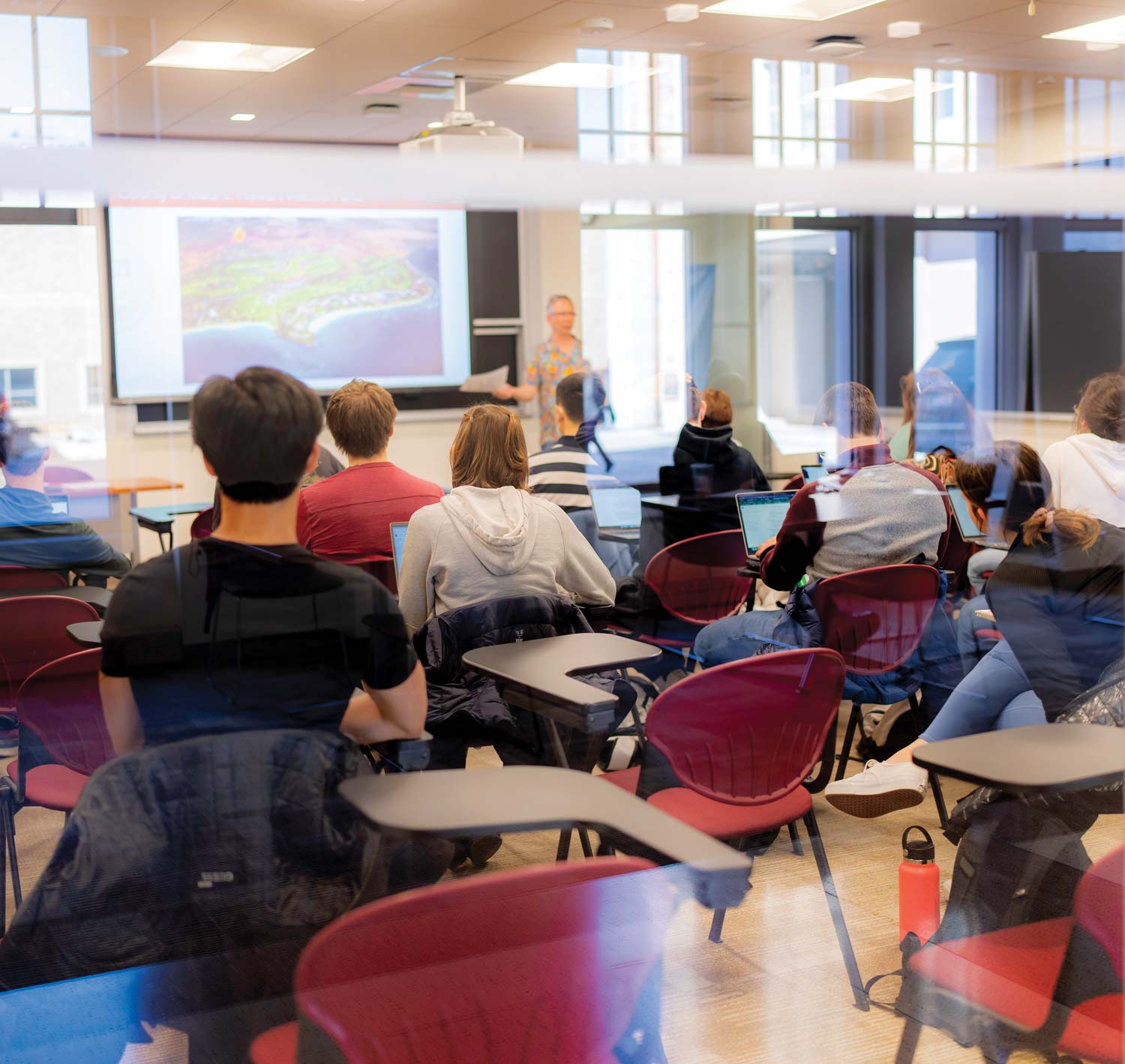 Students sit in a classroom striking the match of curiosity as they face a teacher near a projector screen. Laptops and notebooks are on desks. The room has red chairs and large windows, with reflections visible on the glass.
