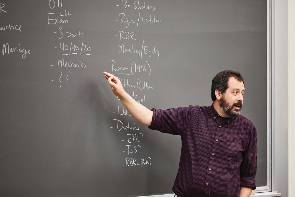 A man with a beard, wearing a maroon shirt, is pointing at a chalkboard covered in handwritten notes and lists in a classroom setting, as if he's about to begin striking the match of inspiration for his students.