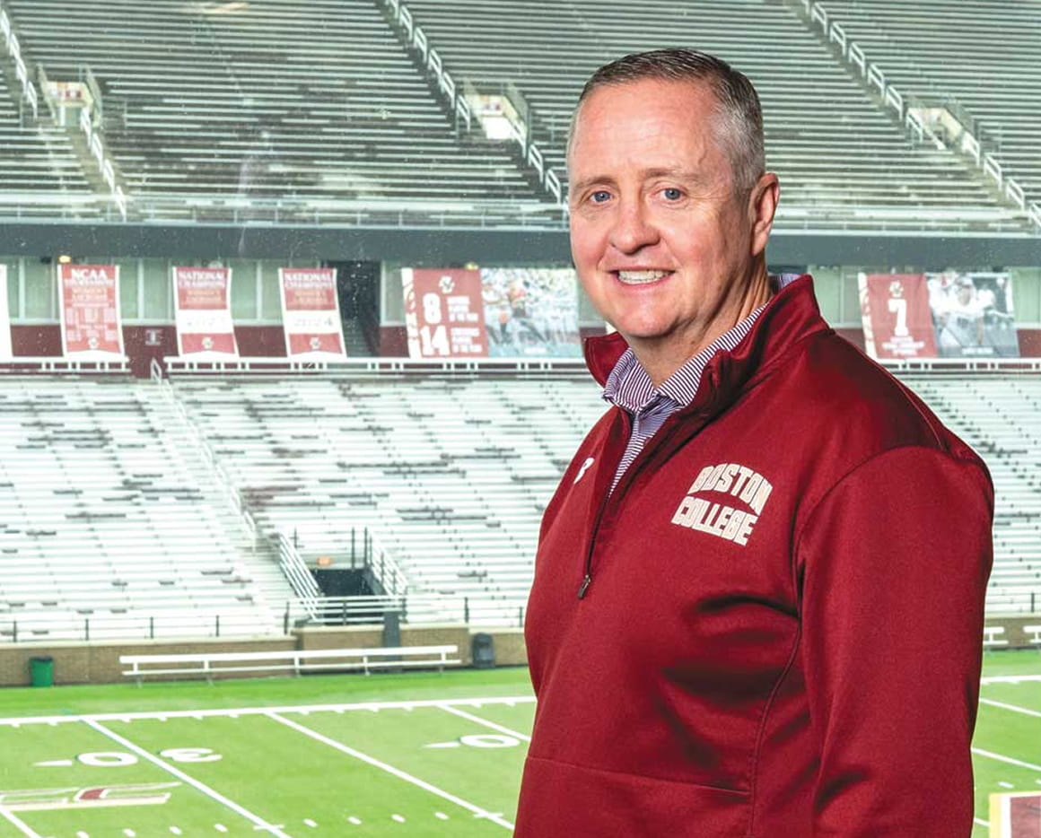 A man in a Boston College jacket stands smiling in front of empty stadium bleachers and a football field.