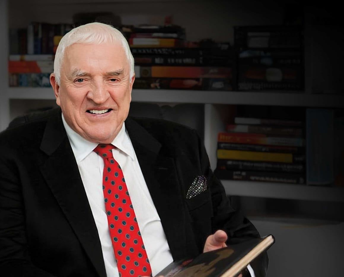 An older man with white hair in a black suit, white shirt, and red polka dot tie sits in front of bookshelves, smiling and holding an open book.